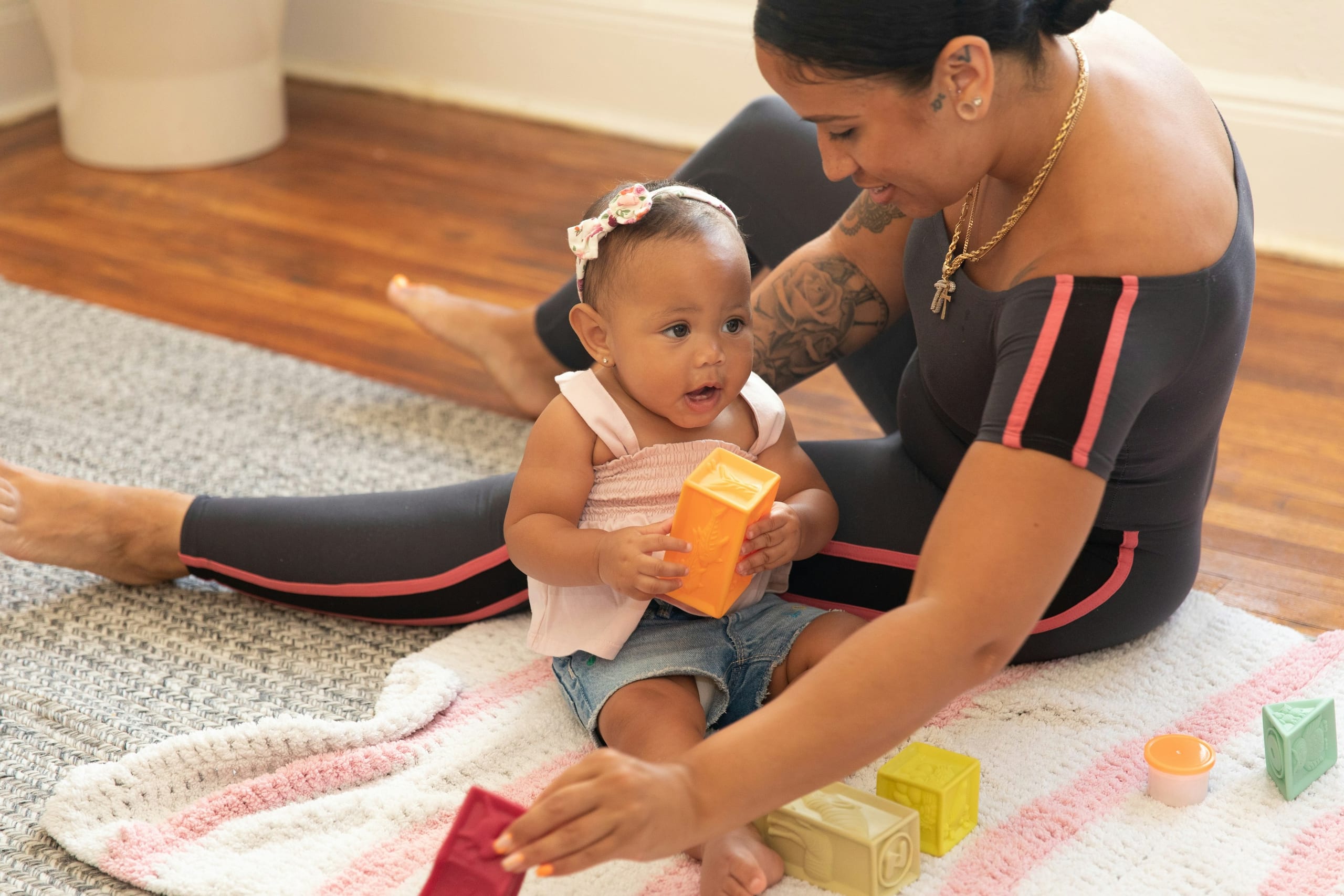 A mother sits on the living room floor beside her baby girl, who holds up an orange block.